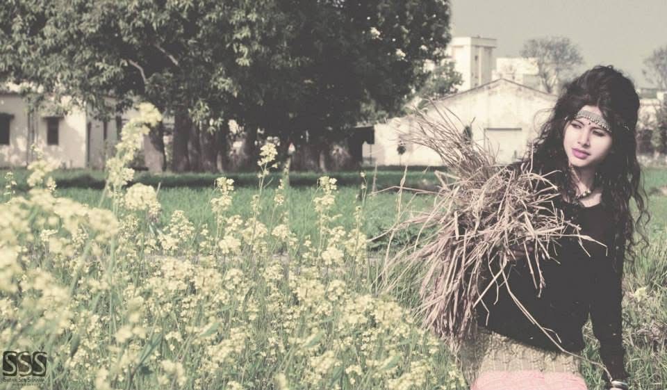 Woman wearing a black hat and a red patterned dress stands outdoors near lush green trees and plants.